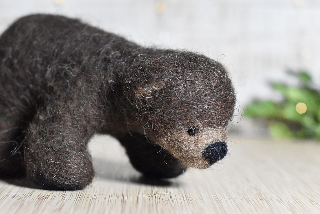 A needle felted wool bear pictured in front of a green branch with bokeh light behind.