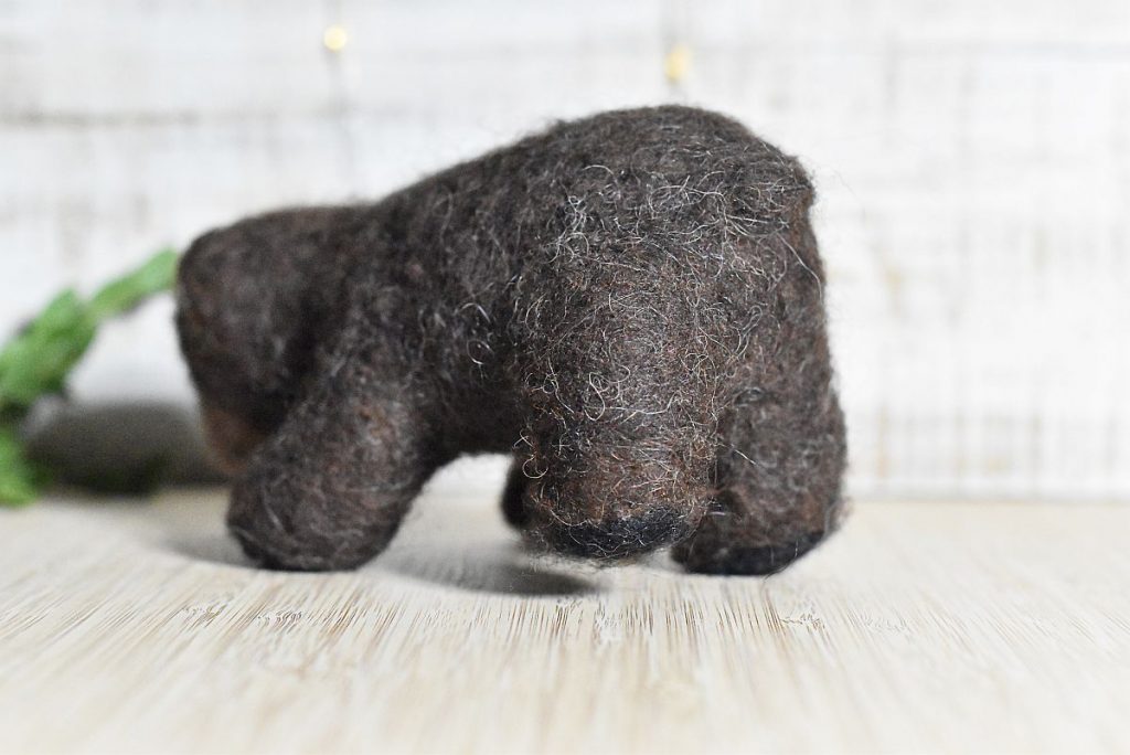 A needle felted wool bear pictured from the back in front of a green branch with bokeh light behind.