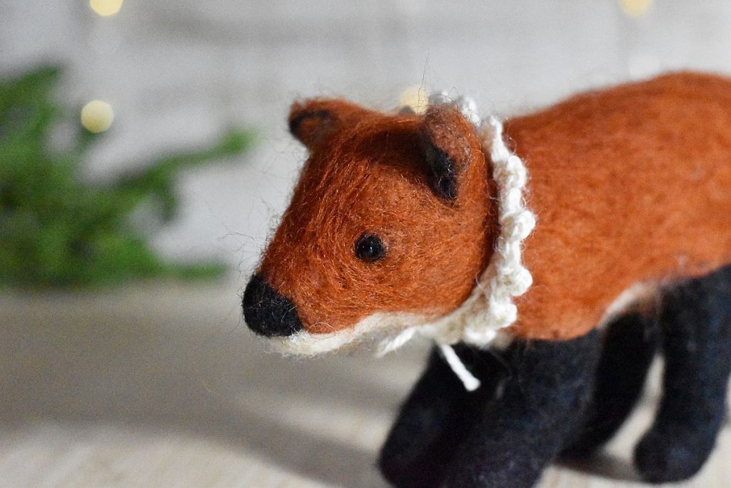 A wool needle felted fox in front of a green branch with bokeh light behind. The fox wears a crocheted collar made from white wool.