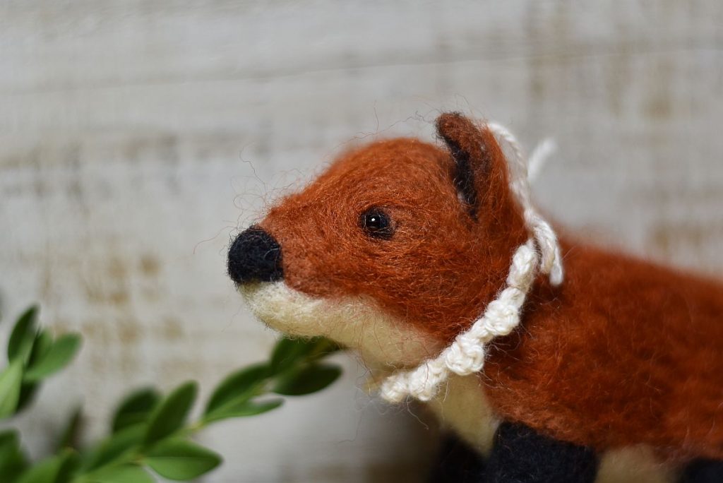 A wool needle felted fox in front of a green branch. The fox wears a crocheted collar made from white wool.