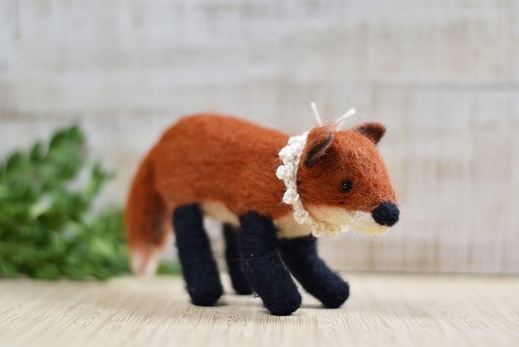 A wool needle felted fox in front of a green branch. The fox wears a crocheted collar made from white wool.