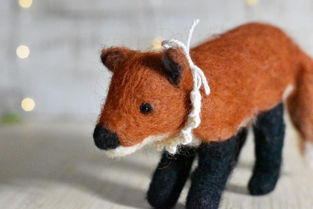 A wool needle felted fox stands on a light wood background with bokeh light behind. The fox wears a crocheted collar made from white wool.