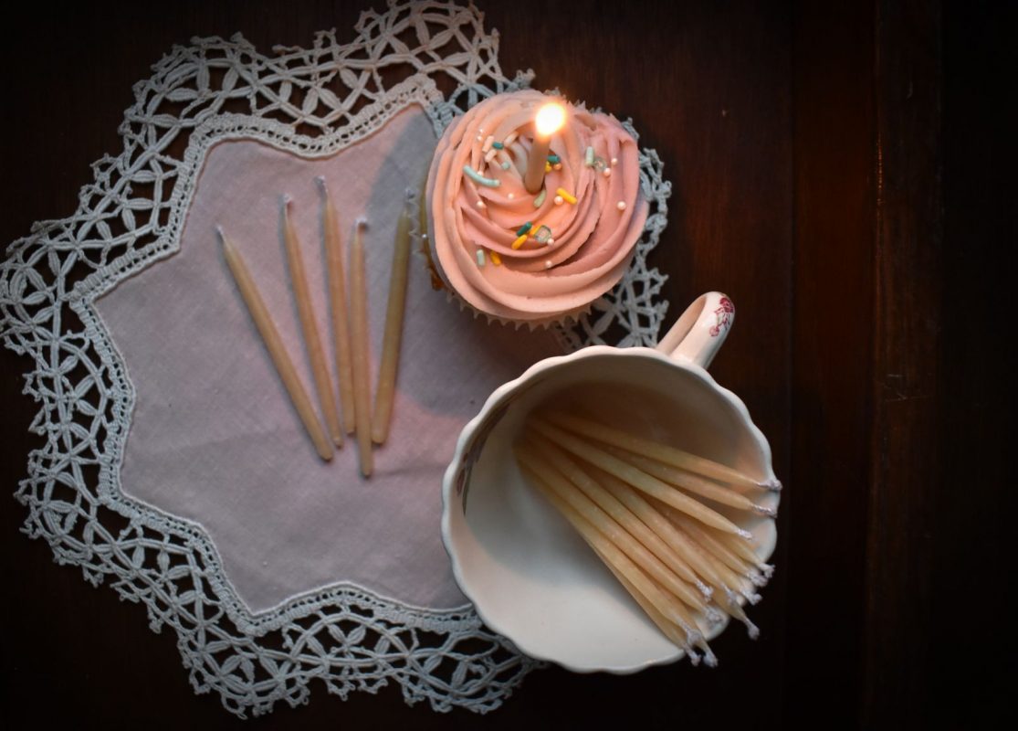 A pink frosted cupcake with a lit candle sits on a vintage napkin beside a cup of beeswax candles