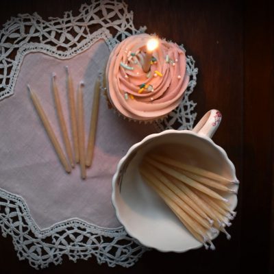 A pink frosted cupcake with a lit candle sits on a vintage napkin beside a cup of beeswax candles
