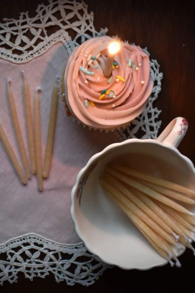 A pink frosted cupcake with a lit candle sits on a vintage napkin beside a cup of beeswax candles