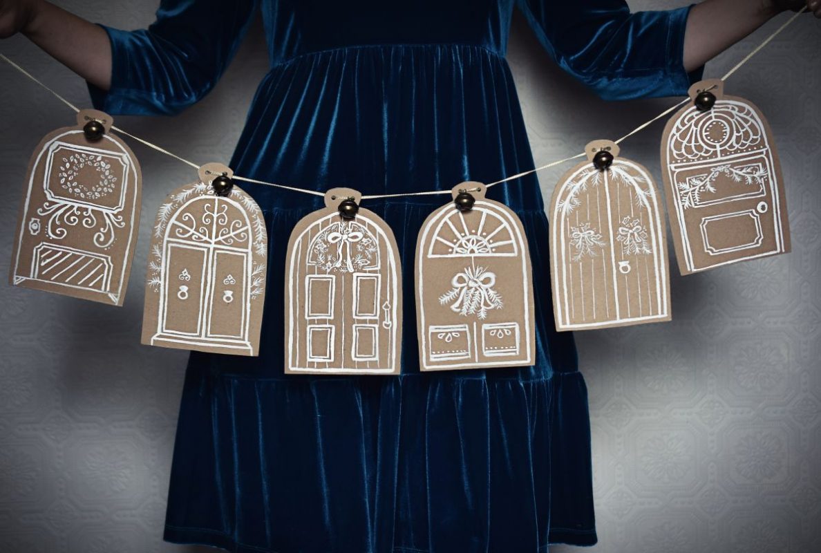 Woman in festive dress holding a hand painted Christmas garland featuring festive doors painted on brown cardboard with white paint