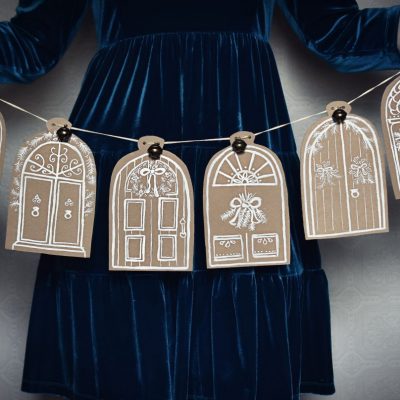 Woman in festive dress holding a hand painted Christmas garland featuring festive doors painted on brown cardboard with white paint