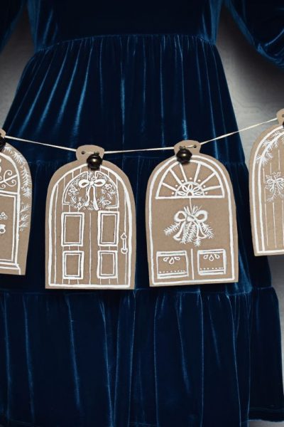 Woman in festive dress holding a hand painted Christmas garland featuring festive doors painted on brown cardboard with white paint